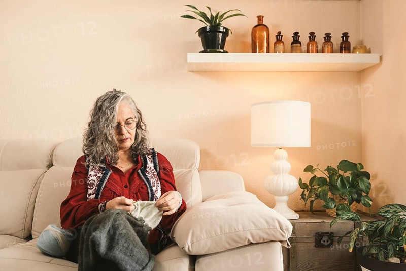 A woman with glasses knits comfortably on a sofa in a warm living room. The space is decorated with a lamp and indoor plants, creating a peaceful ambiance