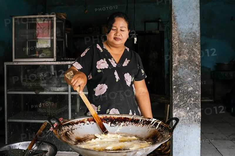 A woman expertly fries food in a large wok, set in a rustic kitchen. Her floral dress adds a touch of color to the scene, capturing traditional cooking methods