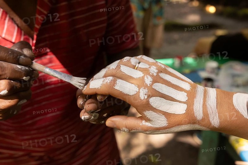 A person paints intricate white patterns on another's hand, capturing the cultural essence of Waigeo Island, Indonesia. The bright colors and cultural involvement are prominent