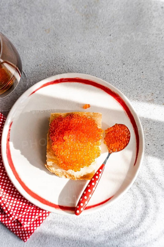 Top view of a vibrant, enticing plate of organic red caviar from trout, served on toast with a red polka dot spoon, against a textured gray background