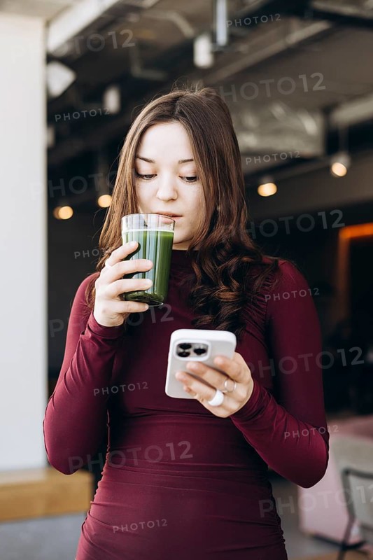 A young female student in a burgundy top enjoys a green smoothie while scrolling through her smartphone in a modern, well-lit setting with an industrial design