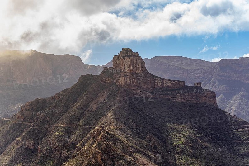 Scenic shot capturing the grandeur of Roque Bentayga under the glowing sunlight, with dramatic clouds above and rugged terrain around, in Artenara, Gran Canaria