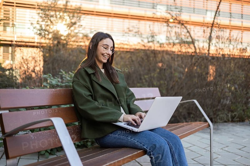A woman in a green jacket and blue jeans sits on a wooden park bench, smiling as she types on her laptop The setting sun casts a warm glow on the urban background