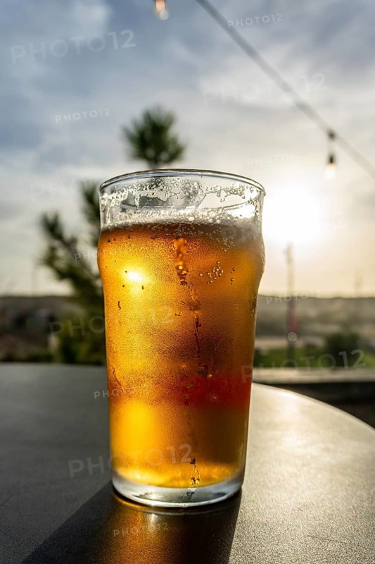 Cold glass of beer with condensation and bubbles, reflecting the warm golden hour light on an outdoor table, evoking summer relaxation and holiday refreshment
