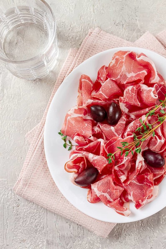Freshly cut dried meat, served with olives and herbs, on a white plate, light background, no people