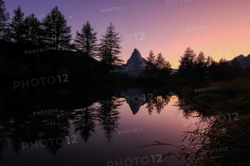 A breathtaking view of the Matterhorn at dawn, captured in the Swiss Alps. Reflecting in a tranquil lake, the iconic peak is silhouetted against a vibrant sky, creating a serene landscape