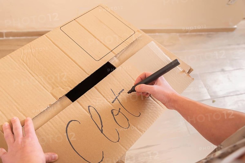 A young woman uses a marker to label a cardboard box with the word Clothes. This image captures the organization and preparation involved in moving a home