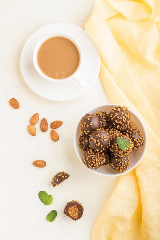 Chocolate caramel ball candies with almonds and a cup of coffee on a white wooden background and yellow textile. top view, flat lay, close up