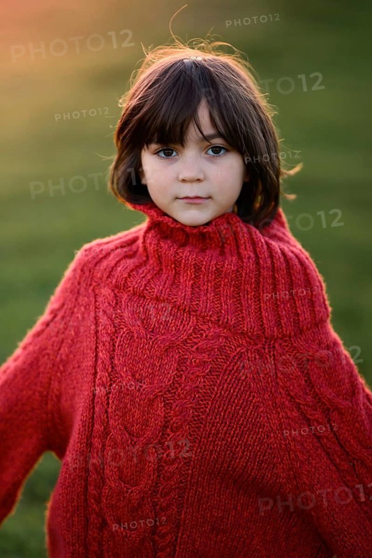 A young girl, dressed in a cozy red sweater, looks thoughtfully at the camera against a background of a golden autumn sunset. She stands in a field symbolizing the warmth and tranquility of the season