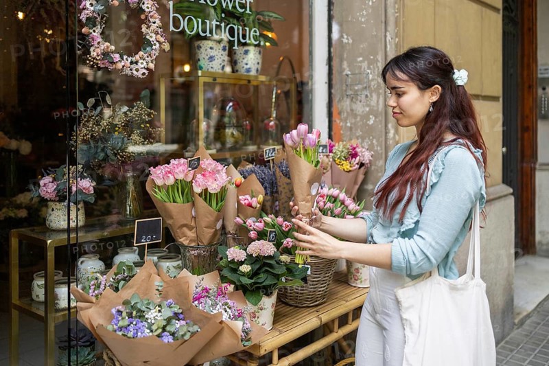 A woman admires assorted bouquets displayed outside a charming flower shop. Her focus on the delicate arrangements highlights a serene moment in an urban setting