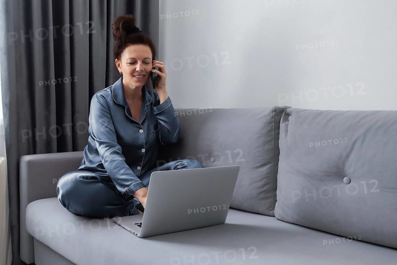 Woman in pajamas sitting on a gray sofa, multitasking with a phone and laptop. She is engaged in remote work or conversation in a cozy, home environment