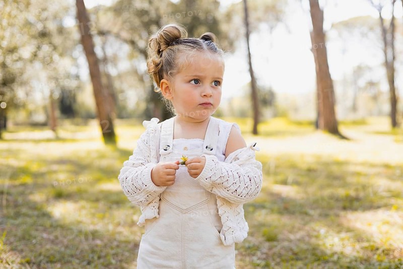 A little girl in a light outfit standing in a sunlit forest, holding a small flower. The serene background, with green trees and grass, highlights the peaceful outdoor setting