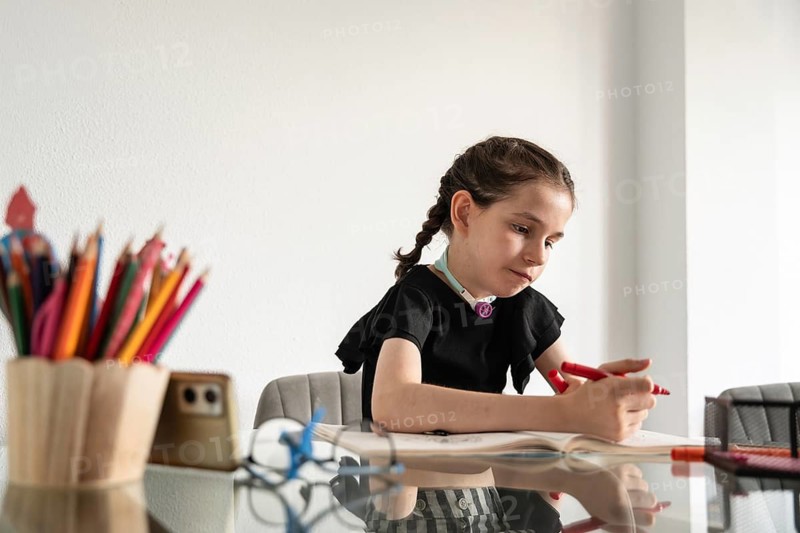 A young girl with a tracheostomy is deeply engaged in drawing at a bright, indoor table. The scene emphasizes her concentration and creativity with colored pencils