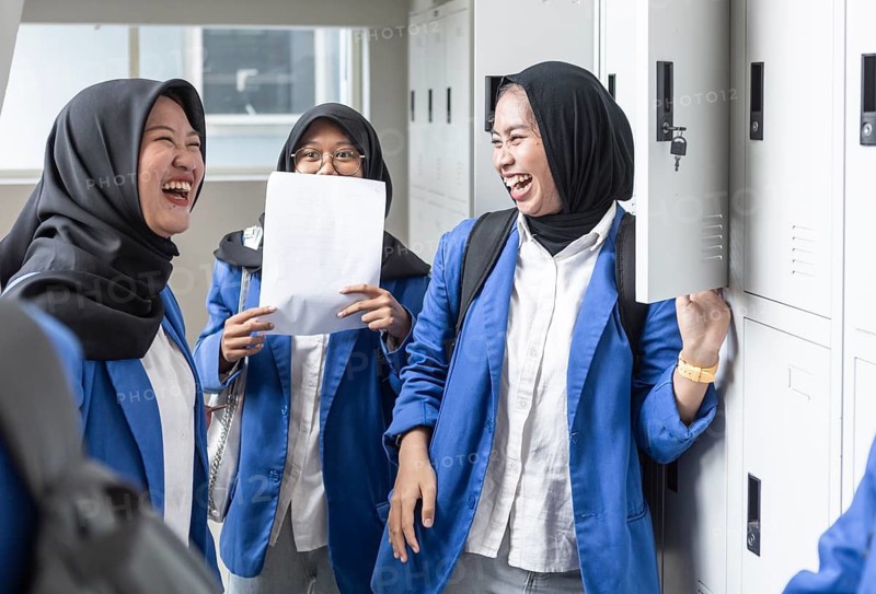 A group of Asian Muslim students in a school corridor, laughing and sharing a moment of camaraderie. They wear blue uniforms and carry backpacks, ready for a new day of learning