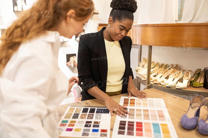 Two women engaged in a color selection task in a fashion boutique, surrounded by shoes. They review color swatches, emphasizing the interplay of fashion and style in design choices