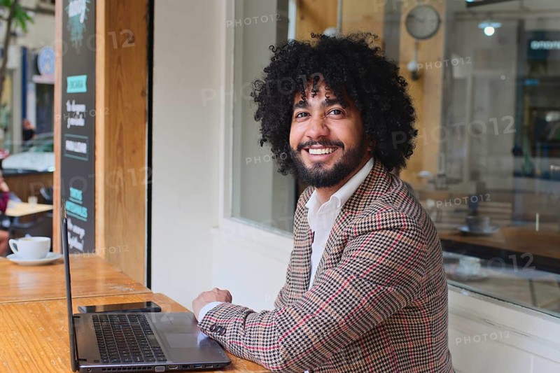 A mixed-race insurance salesman with afro hair smiles at a wooden table, working on a laptop. The indoor setting includes a cafe with warm lighting and white walls