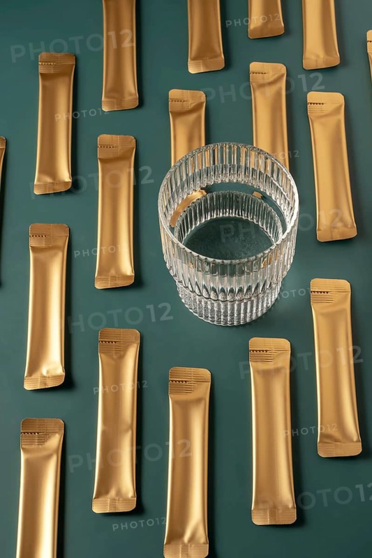 From above a collection of golden jelly collagen sachets next to a glass cup on a dark green background. The elegant and minimalist arrangement highlights the product's appeal