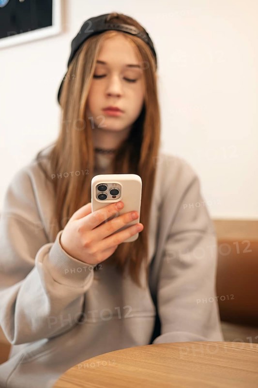 Teenage girl sitting indoors, focused on her smartphone. She holds the device with both hands, wearing a casual hoodie and cap. The scene suggests connectivity and modern technology