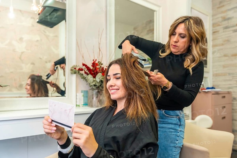 Professional hairstylist straightening client's long brown hair in a modern beauty salon, focusing on hair care and transformation, with the client smiling and checking her reflection