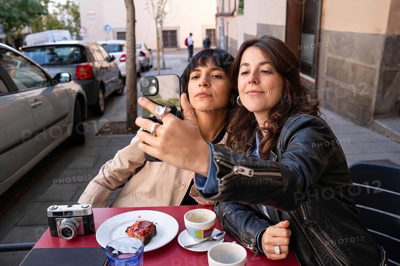 A joyful multiethnic lesbian couple captures a selfie while having coffee outdoors. Celebrating love and diversity in an urban setting, highlighting LGBTQIA+ pride