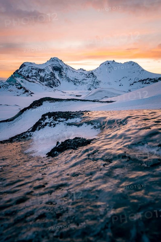 A stunning winter sunset illuminates the snowy ridges and frozen textures of Katla Glacier in Iceland, with vivid sky colors casting a warm glow over the rugged, icy landscape