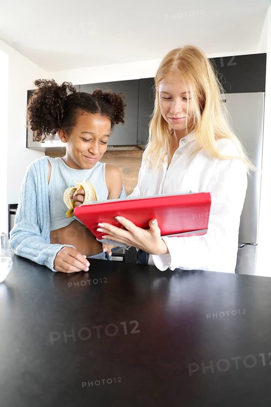 A young woman and a girl stand in a modern kitchen, smiling and enjoying time together. The woman holds a red tablet while the girl eats a banana, engaging in a fun, casual conversation