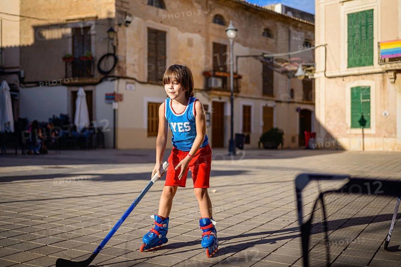 A young child enjoys a game of street hockey, wearing inline skates and colorful sportswear, holding a hockey stick on a sunlit urban street. The scene captures childhood joy and outdoor play