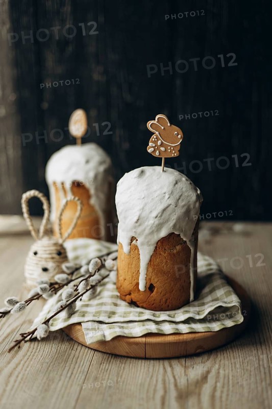 Traditional Russian Easter kulich breads, generously frosted with white icing and adorned with charming Easter themed gingerbread cookies and willow catkin branches displayed on a rustic wooden background