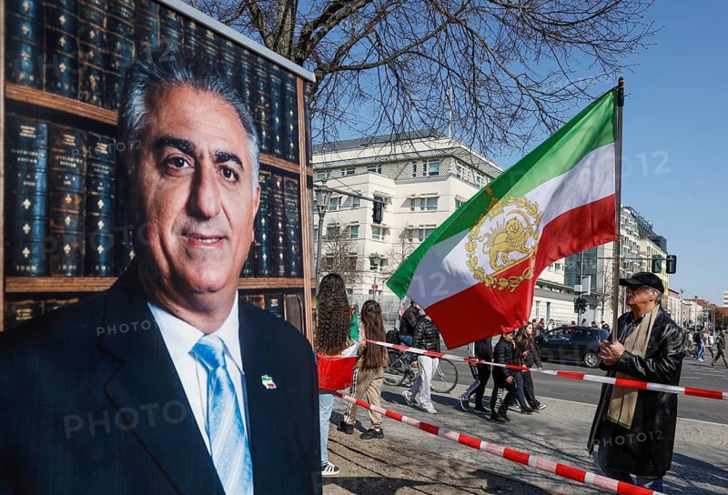 Iran demo for the end of the Islamist mullah regime in Iran, a man standing with a large historic Persian flag with lion and sword at a picture of Prince Reza Pahlavi at the American Embassy in Berlin, 11.04.2026, Berlin, Berlin, Germany