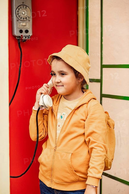 A preteen girl wearing a yellow hat and hoodie casually converses on a vintage phone mounted on a red wall