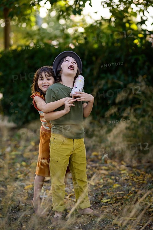 Two children playing outside on a sunny day. One child wearing a hat holds a plush toy while the other embraces them from behind, both smiling in a joyful moment