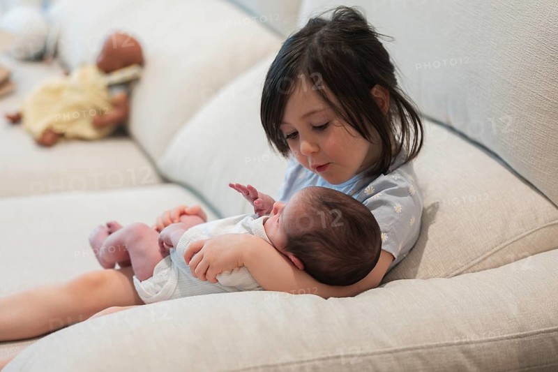 A joyful young family moment as a big sister tenderly holds her newborn sibling on a cozy sofa. The scene radiates warmth, love, and familial bonding