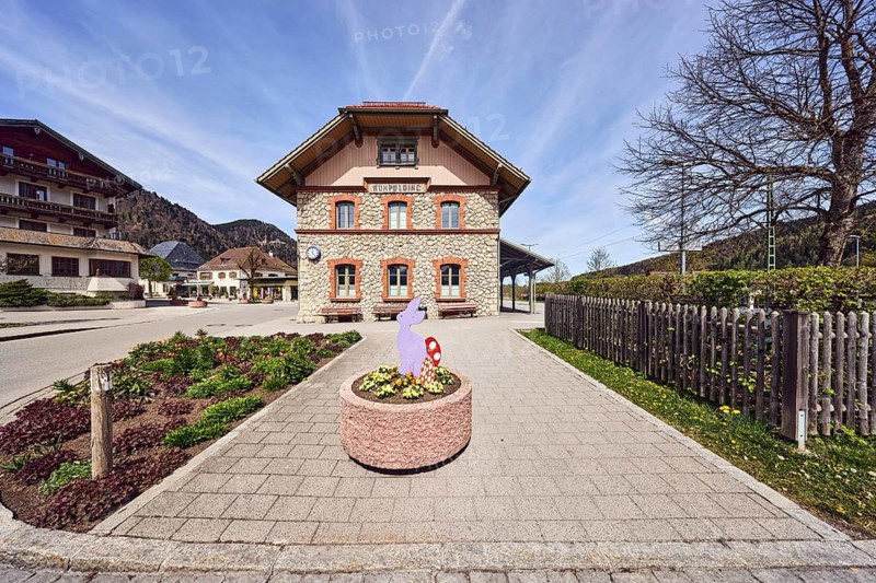 Ruhpolding railway station, public transport, alpine building, commercial building, crippled hipped roof, general architecture, wooden fence, flower bed, flower pot, Easter decoration, benches, hilly landscape, coniferous forest, blue sky, cirrus clouds, cirrostratus clouds, Bahnhofstraße, Ruhpolding, district of Traunstein, Bavaria, Germany