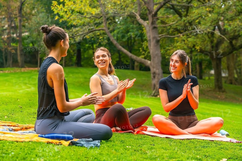 Women friends sitting on yoga mats, clapping hands and smiling happily, sharing a moment of wellness, unity, and support during an outdoor fitness session