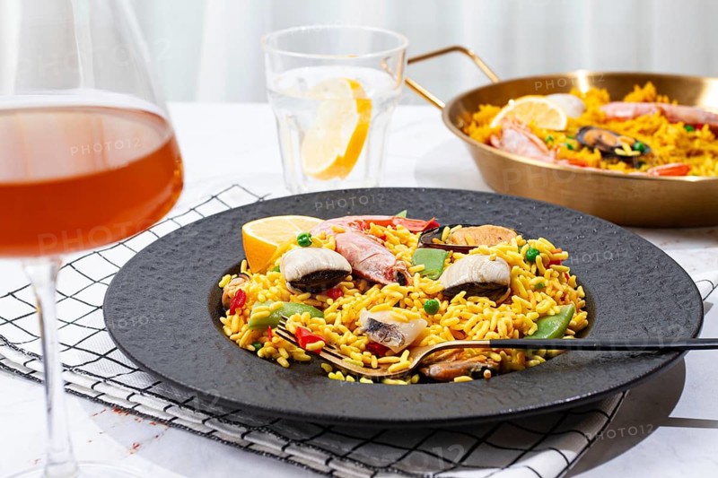 A table is set with a large black plate of seafood paella next to a golden pan. A glass with lemon slices sits nearby, along with another drink. Natural light fills the space