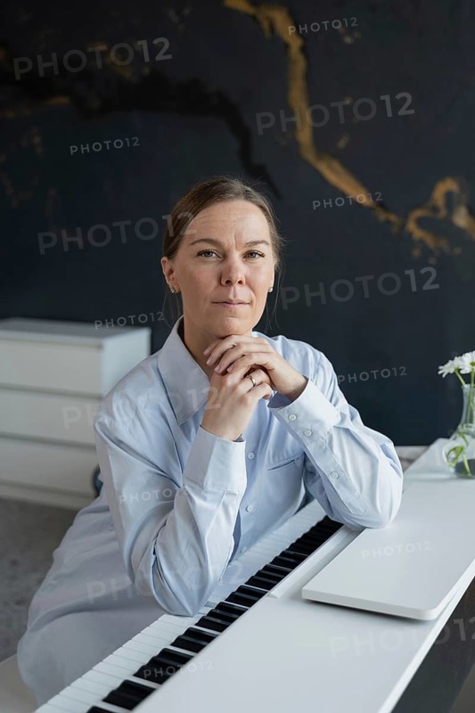 A poised woman in a light blue shirt sits at a piano, looking confidently at the camera. The setting is modern with artistic decor