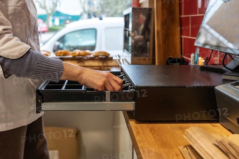 Baker or shop assistant in an apron opening the cash register drawer, preparing to handle money transactions at the sales counter in a bakery or retail store