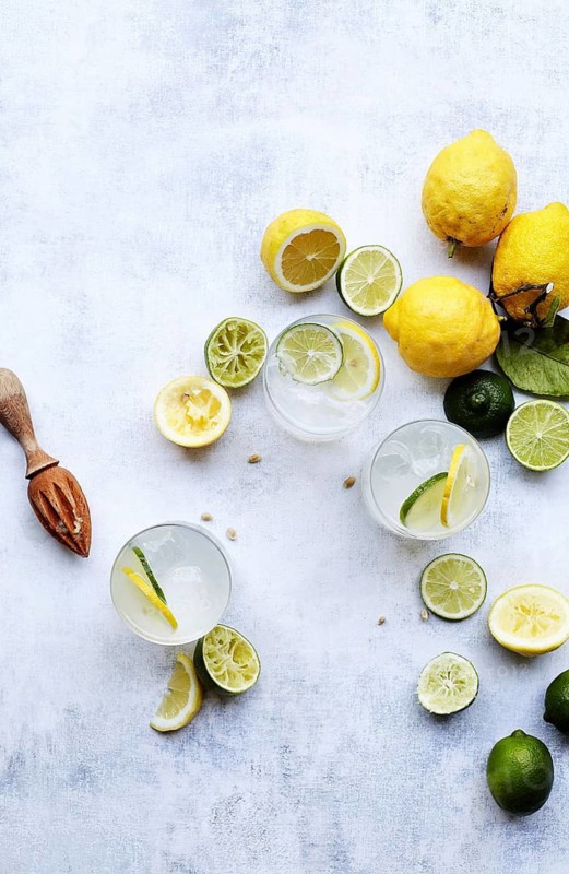 Top view of refreshing summer drinks with slices of lemon and lime on a textured white background. Wood citrus squeezer and scattered seeds add rustic charm