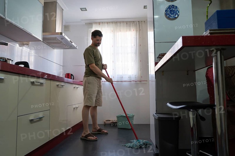 A man is seen mopping the floor in a modern kitchen, reflecting a typical household chore. He is wearing casual attire and appears focused on cleaning the tiled floor