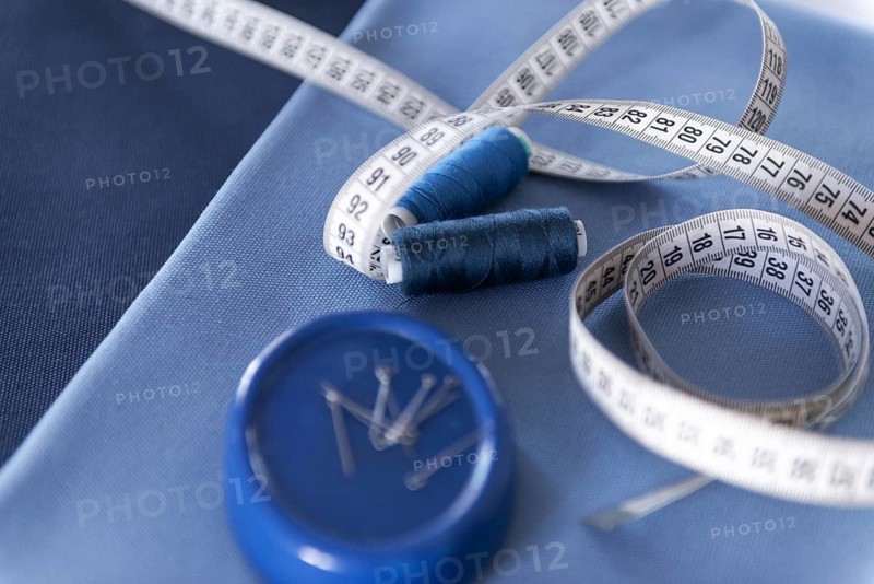 A close-up view of sewing tools including a white measuring tape, two blue thread spools, and a pin holder placed on blue fabrics, representing a sewing workshop environment