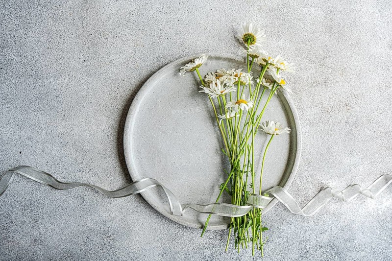 Top view of a bunch of fresh daisy flowers on a round ceramic plate. A graceful ribbon lies on a textured concrete background, creating a serene spring scene