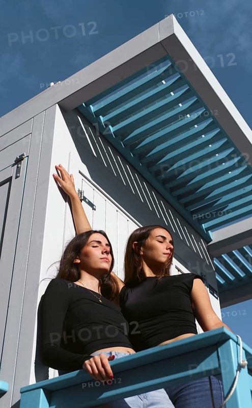 Two women friends enjoy a sunny day together, standing outdoors against a blue sky backdrop. They express friendship and joy in a moment of leisure and relaxation