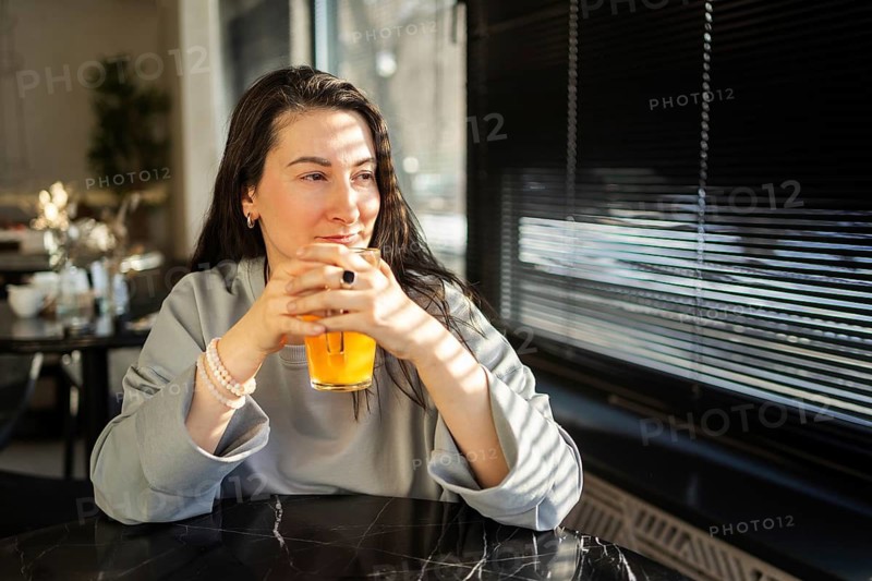 A mid-aged woman gazes out a window while sipping a drink in a sunlit, embodying a moment of peaceful contemplation