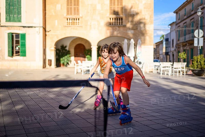 Two children play street hockey on roller skates in a sunlit plaza, showcasing teamwork and energy. The lively scene is set against a backdrop of historic buildings