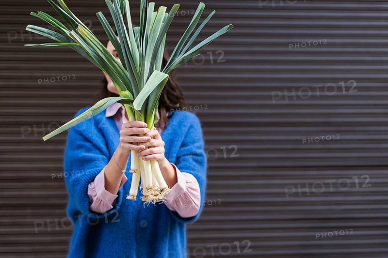 A woman in a blue sweater holds a bundle of fresh vegetables, hiding her face, against a blurred urban backdrop. The leeks green and white contrast her outfit