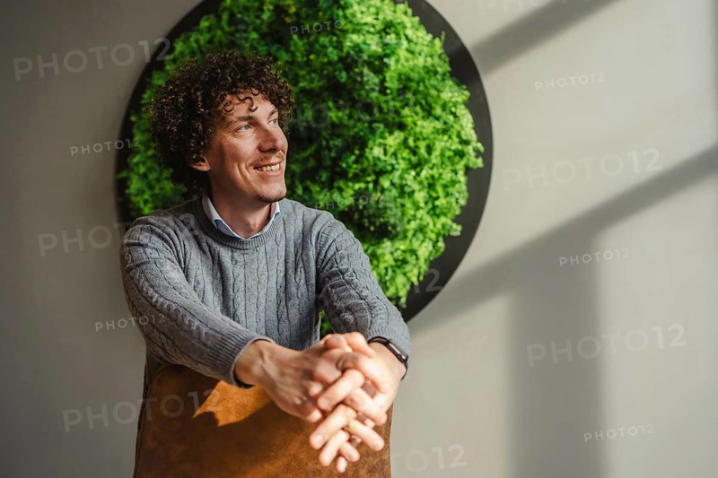 A young curly-haired man smiles while sitting in front of a vibrant green wall, symbolizing themes of ecology and sustainable development, highlighted by natural light