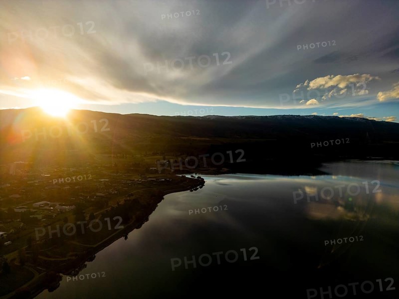 Aerial view of Queenstown in autumn, showcasing a breathtaking sunset over a tranquil lake, with sun rays illuminating the serene landscape surrounded by distant hills