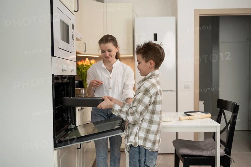 A mother and her young son happily bake in the kitchen. The boy places a tray in the oven as they enjoy quality time and learn new skills