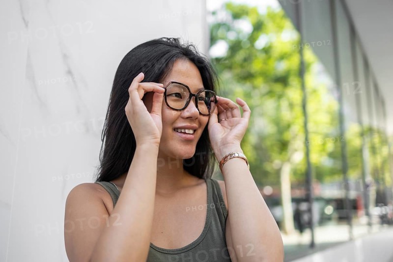 A stylish Chinese woman adjusts her glasses while enjoying a sunny summer day in the bustling city. Her casual attire and joyful expression reflect the vibrant urban life