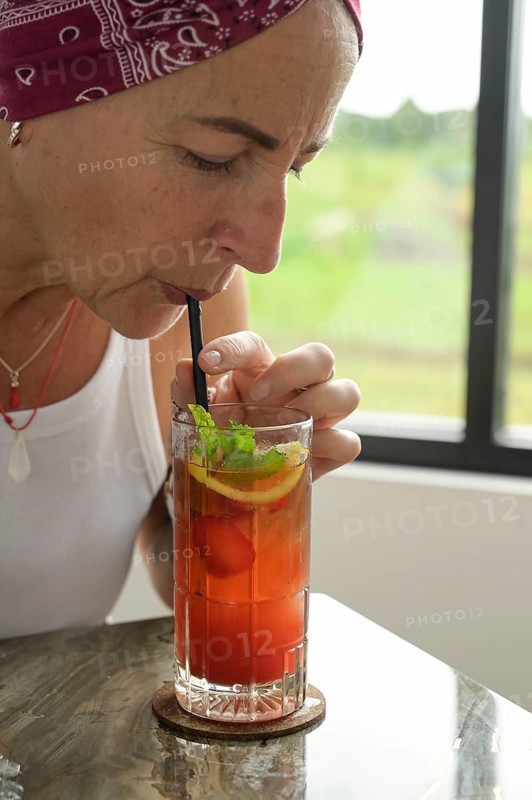 A mature woman with a red headscarf is pictured sipping a colorful Es Teh Nusantara tea drink through a straw. She appears engaged and serene as she tastes the beverage, perfectly accessorized with a sprig of mint